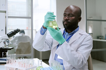 Middle aged Black man wearing lab coat and gloves examining flask with yellow liquid in laboratory, sitting at table with test tubes and microscope, conducting scientific research