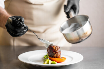 A professional chef wearing black gloves carefully plates a gourmet meal, adding a creamy sauce to a perfectly cooked piece of meat on a white plate in a restaurant kitchen setting.