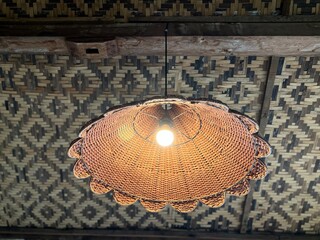 A woven, bell-shaped lampshade with a scalloped bottom edge, illuminated from the center by a warm light bulb, hanging from a ceiling made of dark wood beam and a patterned, woven bamboo matting.