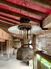 An old, weathered hurricane lantern, also known as a Petromax lamp, hanging from a wooden structure. It has metallic base, a glass, globe, and a white enamel top reflector.