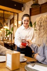 Vertical photo of a young girl with Down syndrome working in a modern cafe wearing a white uniform.The girl is taking a payment from a customer using a card reader while looking at her.