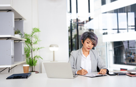 Asian Business woman planning, taking notes and writing in a paper while reading a report, document or paperwork in an office alone at work. One serious corporate professional working at a desk. - Powered by Adobe