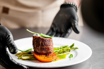 A chef wearing black gloves presents a perfectly cooked, gourmet beef filet mignon dish with asparagus and roasted vegetables on a white plate in a restaurant setting