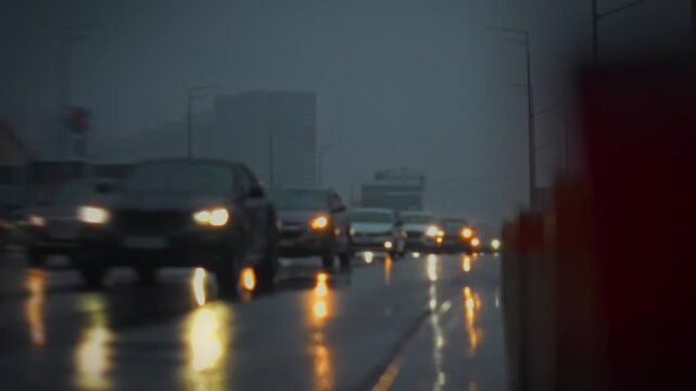 Vehicles drive slowly along a wet highway during a heavy rainstorm in the evening. Reflections of lights can be seen on the road, creating a hazy and moody atmosphere