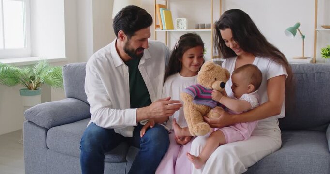 Family parents children bonding and play with toy teddy. Happy mother cuddles baby as father and daughter laugh on sofa at home, sharing warmth, gentle parenting, sibling love, and everyday joy.
