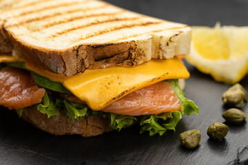 Grilled salmon and cheddar sandwich with lettuce on slate board, lemon and capers
