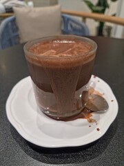 A glass mug of dark hot chocolate or cocoa, served on a white saucer with spilled drops, in a cafe setting with a blurred background.