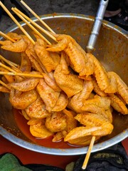 A large metal bowl filled with raw chicken wings that are seasoned and marinated in a bright orange-red sauce, skewered with wooden sticks, ready for grilling or frying.