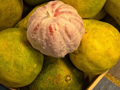 A close-up of a peeled pink pomelo with white pith surrounding the flesh, resting on a pile of unpeeled green and yellow pomelos.