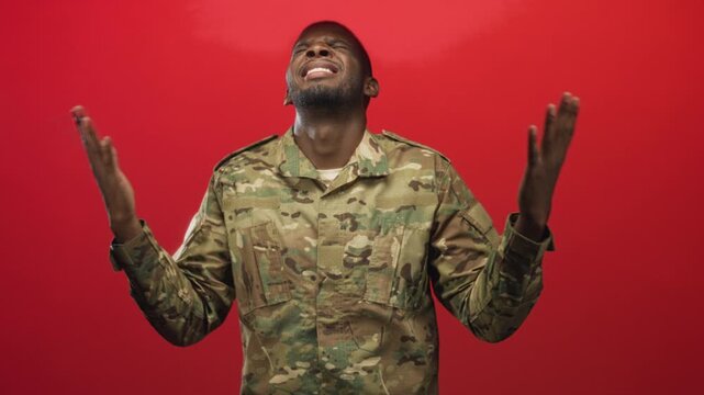 Soldier man in camouflage uniform raises hands and looks upward against a solid red studio backdrop; hope.