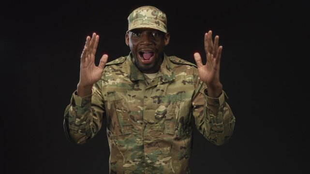 Man soldier in camouflage uniform and cap raises hands with open mouth shouting, isolated on a black studio backdrop; surprise alert.