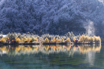 Bipenggou in autumn winter. Snowy mountain forest reflected in clear alpine lake with golden autumn trees and soft morning mist.