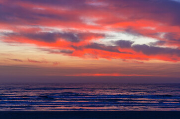 A vibrant ocean sunset with dramatic red, orange, and purple clouds. The deep blue and purple hues of the water below reflect the vivid sky, creating a serene and colorful natural scene