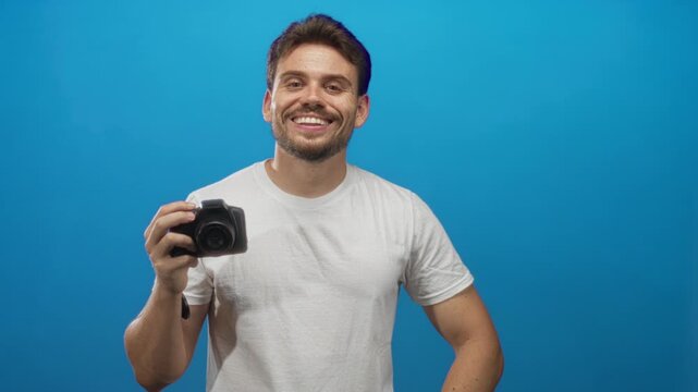 Man holding dslr camera with right hand and left on hip, posing in blue studio; playful confidence casual.