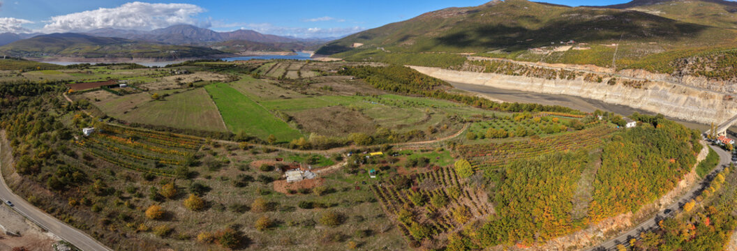 A wide aerial panorama of Kukës showing green fields, orchards and rolling hills near the Drin Valley, with distant mountains and the lake in view. - Powered by Adobe