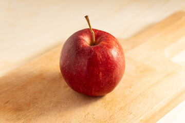 Single Red Apple with Stem Resting on a Light Wooden Cutting Board in Soft, Warm Light
