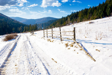 winter countryside in mountains on a sunny day. wooden fence along the road through coniferous forest near snow covered hills. scenic view of rustic scene in western ukraine. carpathian wonderland