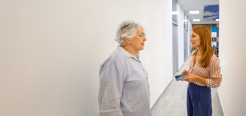 Elderly patient discussing medical concerns with female doctor holding smartphone in hospital corridor by white wall