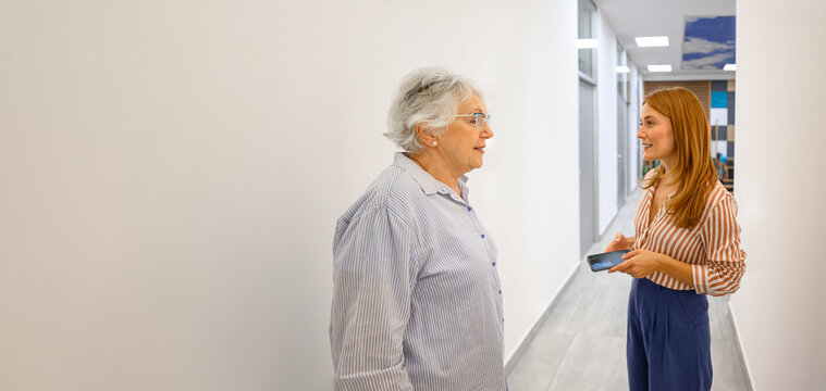 Elderly patient discussing medical concerns with female doctor holding smartphone in hospital corridor by white wall
