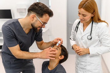 Obraz premium Female doctor looking at coworker putting eye drops in boy's eye during hospital visit at ophthalmology clinic