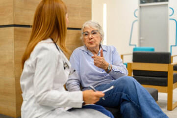 Obraz premium Senior woman discussing symptoms with female doctor taking notes on clipboard in hospital during routine checkup