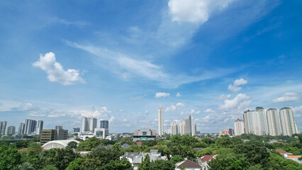 Jakarta city skyline under bright blue sky with scattered clouds, featuring modern high-rise buildings and lush green residential areas. © Rijjal