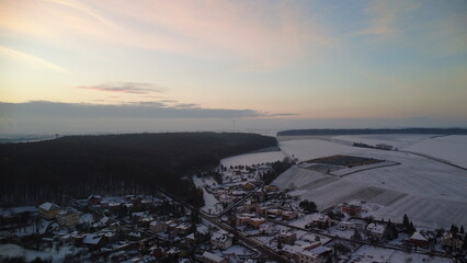 Sunset over a snow-covered Czech landscape, featuring rolling fields, trees, and a serene winter atmosphere. The warm colors of the setting sun contrast beautifully with the fresh snow.