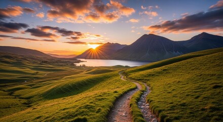 A winding hiking trail meanders through rolling green hills towards a majestic mountain lake during a spectacular golden hour sunset with vibrant sun rays