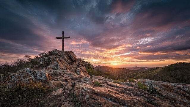 Wooden cross atop rocky hill against a dramatic sunset and cloudy sky