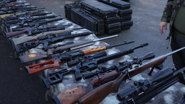 Rifles with scopes arranged in rows on a camo table during an outdoor inspection creating a controlled secure scene focused on examining seized long guns in clear daylight conditions