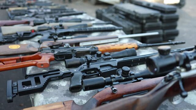 Rifles arranged in rows on a camo table during an outdoor weapons inspection create a focused controlled scene showing seized long guns prepared for detailed examination in a secure daylight environme