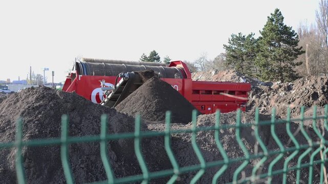 Soil and debris move along a red industrial screening machine forming piles as the rotating drum separates mixed ground materials creating a clear scene of outdoor waste sorting and earth processing o