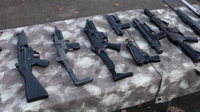 Confiscated submachine guns and pistols on a camo table as officers inspect seized illegal firearms creating a tense atmosphere of arms trafficking field investigation