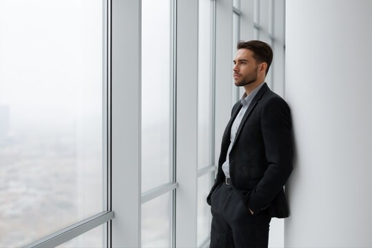 Young caucasian male businessman in formal attire standing by office window contemplating