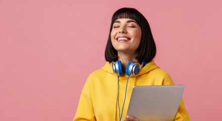 A happy young woman with dark hair in a yellow hoodie smiles with her eyes closed, holding a laptop and wearing blue headphones around her neck, feeling joyful and relaxed
