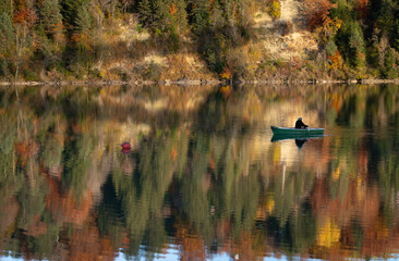 A small rowboat glides across calm waters in autumn. In the background, tall trees are colored with autumn hues and reflected in the water.