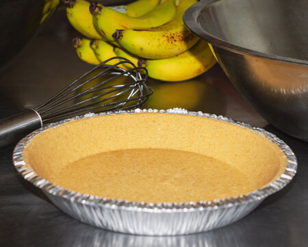 A ready-made graham cracker pie crust on a stainless steel counter next to a whisk and ripe bananas, ready to be assembled into a creamy dessert.