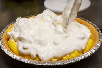 A generous layer of fresh, fluffy whipped cream is being spooned onto the bright yellow banana cream filling of a pie in a commercial kitchen.