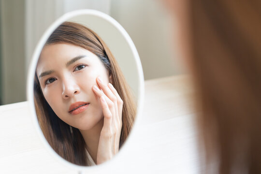 Anti aging, worry puberty asian young woman, stressed girl looking in mirror, allergy presenting an allergic reaction from cosmetic on face. Beauty, health care from acne problem, dry skin, treatment.