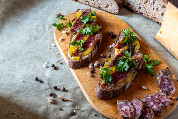 Top view of two gourmet open sandwiches with cured meat and herbs on rustic rye bread, presented on a wooden board alongside sliced salami and a block of cheese