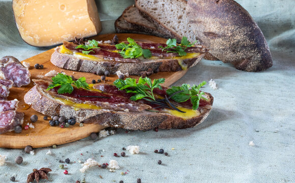 Artisan open-face sandwiches with cured meat and herbs on rustic bread, featuring a block of hard cheese and a loaf of bread in the background on a light linen tablecloth - Powered by Adobe