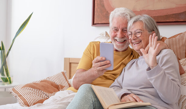 Video call concept. Portrait of happy senior couple lying in bed while using phone for a video call. Serene retired couple