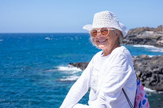Carefree smiling senior woman in white hat looking at camera enjoying a sunny day at the seaside with the blue ocean in the background - Powered by Adobe