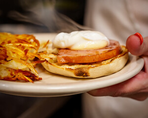 A chef presents a plate with a breakfast sandwich—English muffin, ham, and a poached egg—next to hash browns in the Waukesha cafe.