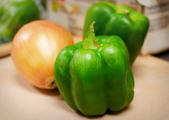A close-up of fresh, whole green bell peppers and a yellow onion resting on a wooden cutting board, ready for chopping in the cafe kitchen.