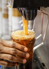 A cafe worker's hand holds a glass being filled with bubbling, dark brown soda from a self-service or fountain beverage machine.