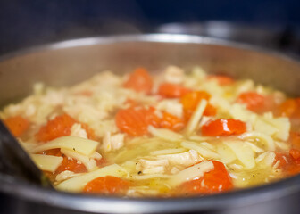 A close-up of hearty, steaming chicken noodle soup, with chunks of chicken and bright carrots, simmering in a pot on a cafe stovetop.