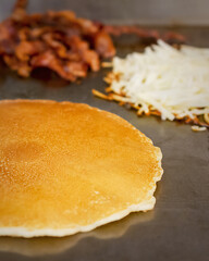 A golden pancake, shredded hash browns, and bacon cooking on a large flat-top griddle in a busy Waukesha cafe kitchen.
