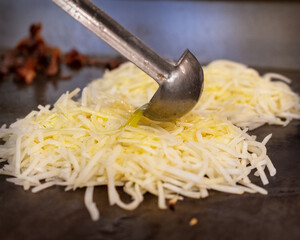 A chef uses a ladle to pour melted butter over shredded hash browns cooking on a hot flat-top griddle, preparing a savory breakfast.