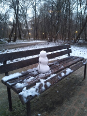 Small Snowman Sitting Alone on a Park Bench in an Early Winter Landscape with Bare Trees at Dusk_1